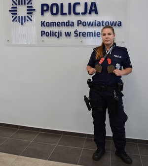 Policjantka w sali konferencyjnej stoi przy logo śremskiej Policji i prezentuje medale